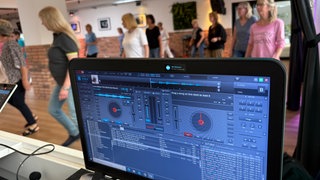 Ein Monitor zeigt abgespielte Musik, im Hintergrund tanzen Menschen