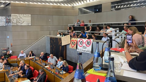 Mehrere Menschen sitzen im Gemeinderat in Pforzheim. Zwei Zuschauerinnen halten drei Plakate hoch, die gegen die Abschaffung der Tempo 30 Zone gerichtet sind. Der Gemeinderat in Pforzheim hat entschieden, dass an vielen Hauptverkehrstraßen Tempo 30 abgeschafft werden soll.