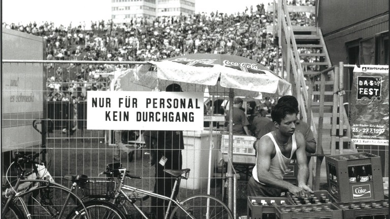 DAS FEST 1997: Ein Mitarbeiter hebt eine Flasche Getränke. Neben ihn steht ein Zaun, auf dem ein Schild mit der Aufschrift "Nur für Personal - Kein Durchgang" angebracht ist.