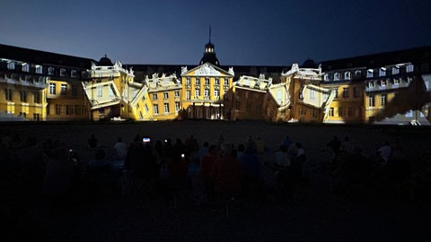 Bei bestem Sommerwetter haben die Schlosslichtspiele Karlsruhe begonnen. Schlossfassade mit Videoinstallation, davor Publikum.