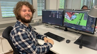 Ein Mann sitzt vor einem Monitor und lacht in die Kamera.