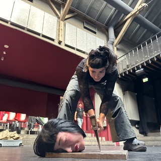 Atoll Festival in Karlsruhe: Ein Puppenkopf liegt auf dem Boden. Die Künstlerin La Vispera steht mit einer Axt davor.