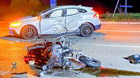 Nach einem Unfall liegt ein beschädigtes Motorrad auf der Straße, dahinter ist ein ebenfalls beschädigtes Auto zu sehen.