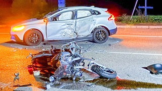 Nach einem Unfall liegt ein beschädigtes Motorrad auf der Straße, dahinter ist ein ebenfalls beschädigtes Auto zu sehen.