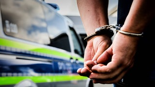 Ein 19-Jähriger soll einen Mann in Wildberg getötet haben. Polizeiauto, davor Hände in Handschellen.