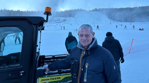 Andreas Kern, der Geschäftsführer des Skilifts Mehliskopf steht vor der Piste. Im Hintergrund fahren Menschen Ski, die Schneekanonen laufen.