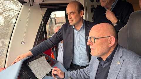 Drei Menschen stehen im Führerhaus der Bahn. 