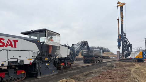 Eine Asphaltfräse entfernt den alten Straßenbelag der A8, während ein Bohrer schon Löcher für die Brücke der Verkehrsschilder in den Boden bohrt.