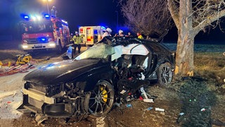 Schwarzes schwer beschädigtes Auto steht neben einem Baum, Einsatzkräfte im Hintergrund