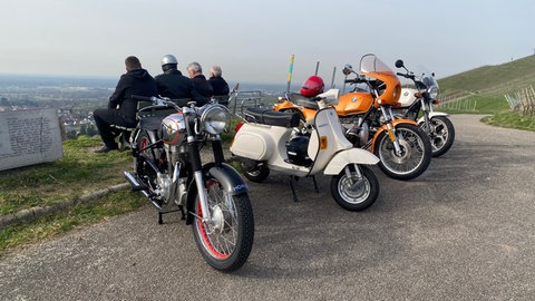 Start der Motorradsaison: Eine Gruppe von Männern sitzt auf einer Bank hinter ihren Oldtimern und schaut in ein Tal.