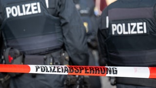 Symbolbild: Razzia - Polizeibeamte stehen bei einer Razzia hinter einem Absperrungsband