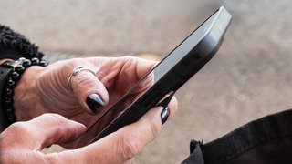 Symbolbild: Eine Frau hält ein Smartphone in der Hand. Die Polizei ermittelt wegen eines Mannes, der unerlaubt Videos von Mitarbeitern in Geschäften gemacht und im Netz veröffentlicht haben soll.