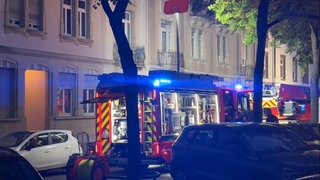 Feuerwehrfahrzeuge stehen vor einem Mehrfamilienhaus in der Karlsruher Weststadt, aus der Eingangstür leuchtet es orange. Das Feuer ist das dritte innerhalb eines halben Jahres im selben Wohnhaus.