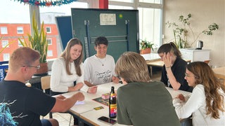 Sechs Studentinnen und Studenten bei ihrem wöchentlichen Spieleabend in Karlsruhe. Sie spielen ein Brettspiel auf einem Tisch.
