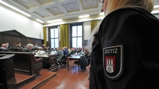 Menschen sitzen in einem Hamburger Gerichtssaal, im Vordergrund eine Justizbeamtin