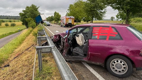 Ein zerstörtes Auto steht an einer Leitplanke. Im Hintegrund liegt ein Sattelzug im Graben, daneben ein Feuerwehrauto. Auf der B293 bei Betten kam es ein Unfall zwischen den beiden Fahrzeugen.