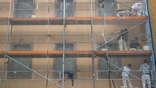 (Symbolbild) Arbeiter auf einem Baugerüst verrichten Arbeiten an der Fassade eines Hauses. Eine Plattform von Forschenden des KIT in Karlsruhe soll bei der Planung für Sanierungen helfen.