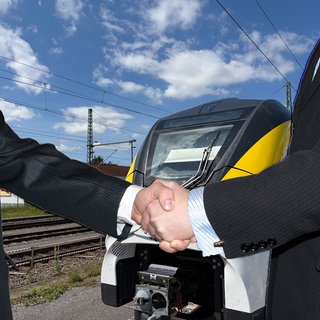 (Symbolbild) Handschlag von zwei Männerhänden vor einem Zug der Hesse-Bahn