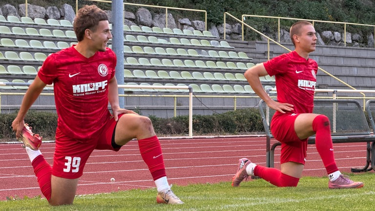 Zwillinge im roten Trikot dehnen sich auf einem Fußballplatz.