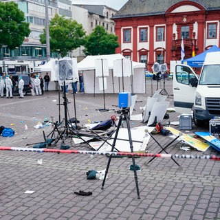 Mitarbeiter der Spurensicherung stehen auf dem Marktplatz hinter einem zertrümmerten Stand. Bei einem Einsatz auf dem Mannheimer Marktplatz hat die Polizei einen Angreifer niedergeschossen. Der Mann wurde dabei verletzt. Er hatte zuvor mehrere Menschen mit einem Messer angegriffen und verletzt, ein Polizist starb am Sonntag an den Folgen.