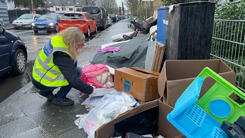 Ein großer Haufen Sperrmüll am Straßenrand. Sabrina Brand trägt eine grelle Warnweste und kniet neben dem Müll. Sie sucht nach Hinweisen.