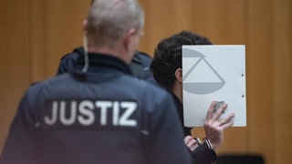 Der angeklagte Sulaiman A. sitzt während eines Prozesstages auf der Anklagebank und hält sich einen Ordner vors Gesicht.