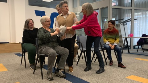 Die Senioren sitzen wie in einem Bus, die Seminarleiterin reißt einer Seniorin die Zeitung aus der Hand.