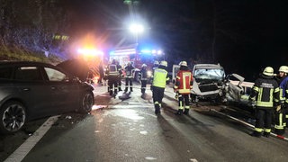 Ein Unfall auf einer Landstraße. Man sieht zwei Autos, die frontal zusammengestoßen sind und Feuerwehr- und Rettungskräfte.
