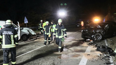 Ein Unfall auf einer Landstraße. Man sieht zwei Autos, die frontal zusammengestoßen sind und Feuerwehr- und Rettungskräfte.