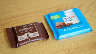 Eine Tafel mit Schokolade der Marke Ritter Sport (r) und ein Haferriegel der Firma Wacker mit der Aufschrift "Monnemer Quadrat Bio" liegen nebeneinander auf einem Tisch. 