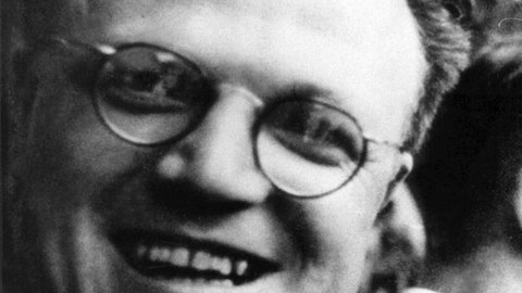 Portraitbild von Alfred Delp. Er trägt eine runde Brille, nach hinten gekämmte Haare und lächelt.