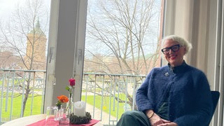 Eine Frau, mit hellen kurzen Haaren und einem freundlichen Lächeln sitzt vor einem Fenster. Vor ihr steht ein kleiner Tisch. Darauf steht eine Kerze, eine Rose und Flyer der TelefonSeelsorge Rhein-Neckar