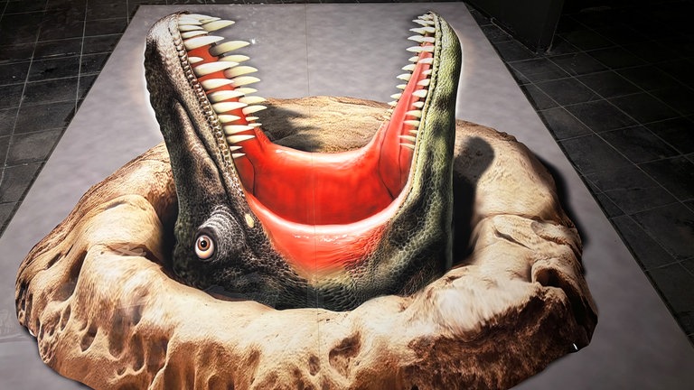 Ein Dinosaurier wirkt so, als ob er aus dem Erdboden herausschaut und sein Maul aufreißt.