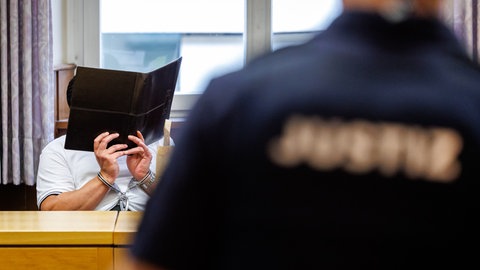 Der Angeklagte hält sich einen Aktenordner vor das Gesicht