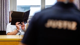 Der Angeklagte hält sich einen Aktenordner vor das Gesicht