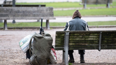 Ein Mann sitzt mit seinen Habseligkeiten auf einer Parkbank.