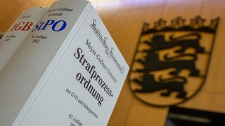 In Bad Überkingen (Kreis Göppingen) und Urbach (Rems-Murr-Kreis) hatte die Polizei vor einigen Monaten Handgranaten gefunden, die mit der Schussserie im Raum Stuttgart zusammenhängen sollen. Vier Männer sind deswegen jetzt angeklagt. Auf dem Bild ist ein Buch mit der Strafprozessordnung vor dem BW-Landeswappen in einem Gerichtssaal zu sehen.