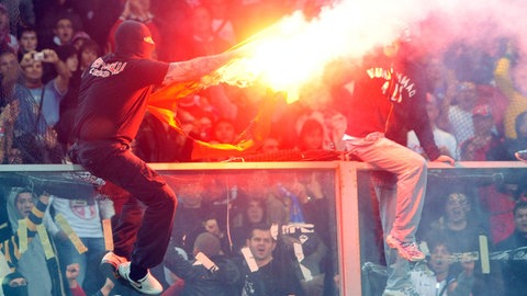 Zwei Fußballfans sitzen vermummt auf einem Zaun und halten einen Bengalo in der Hand (Symbolbild)