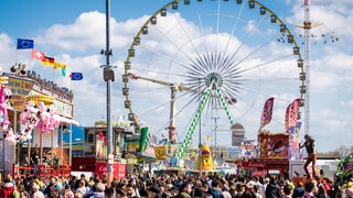 Das Datum vom Frühlingsfest 2024 in Stuttgart auf dem Wasen: 20. April bis 12. Mai. Wer ins Zelt will, sollte einen Tisch reservieren. Der Bierpreis ist gestiegen. Zu sehen sind auf dem Bild Menschenmengen auf dem Festplatz und Fahrgeschäfte, im Hinterhrund steht das Riesenrad.