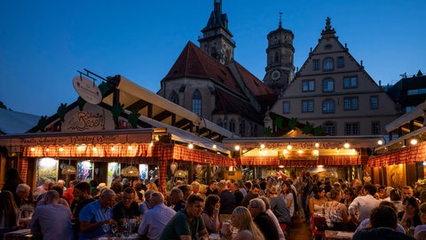 Menschen sitzen auf dem Stuttgarter Weindorf zusammen, trinken Wein und essen schwäbische Spezialitäten. 
