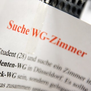  Ein Zettel mit der Aufschrift "Suche WG-Zimmer" hängt an einem Schwarzen Brett.