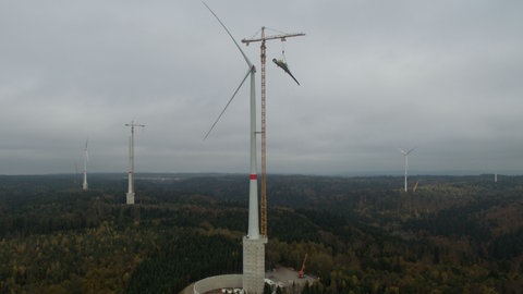 Ein Rotorblatt wird in der Stadt Gaildorf (Baden-Württemberg) an der höchsten Windkraftanlage der Welt an Land von einem Kran hochgezogen (Drohnenaufnahme). Das Projekt kostet mehrere Millionen Euro.