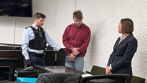 Der Angeklagte im roten Oberteil steht neben seiner Verteidigerin im Saal des 