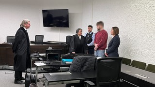 Der Angeklagte im roten Oberteil steht neben seiner Verteidigerin im Saal des 