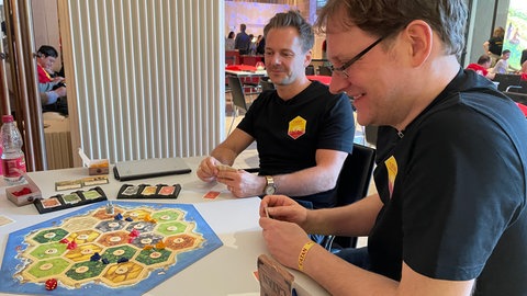 Benjamin Teuber (links) ist der Sohn des kürzlich verstorbenen Erfinders von Catan, Klaus Teuber. Am Rande der Catan-WM 2025 in Stuttgart teste er mit Kosmos-Redaktionsleiter Arndt Fischer (rechts) eine kleine Erweiterung für das Brettspiel.