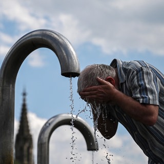 Ein Passant kühlt sich an einem Mineralwasserbrunnen ab, im Hintergrund ein Kirchenturm.