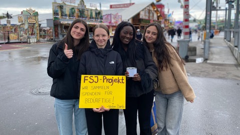 Vier Mädchen auf dem Frühlingsfest. Sie halten ein Schild mit der Aufschrift "Wir sammeln Spenden für Bedürftige!" in der Hand.