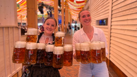 Wasn-Kellnerin Chrissi (li.) und SWR Reporterin Anabel Giesbert tragen Bierkrüge.