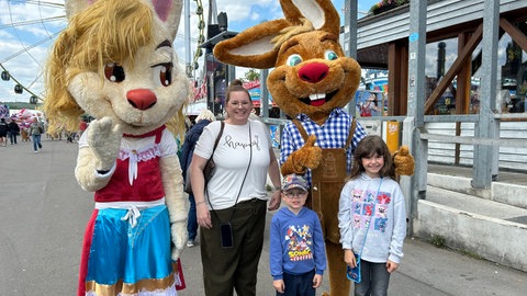 Anja (36) aus Schwieberdingen steht mit ihren beiden Kindern Charlotte (7) und Lennard (4) neben Maskottchen auf dem Stuttgarter Frühlingsfest. 