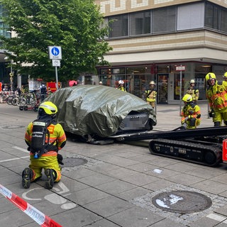 Feuerwehrleute transportieren ein abgedecktes E-Auto nach einem Brand in einer Tiefgarage mit einem speziellen Gerät ab.